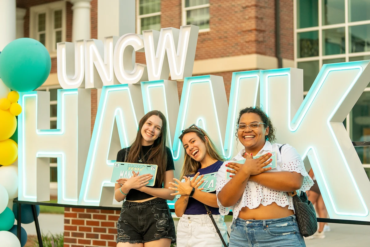 Three people pose happily in front of a large lit-up "UNCW HAWK" sign with colorful balloons nearby.