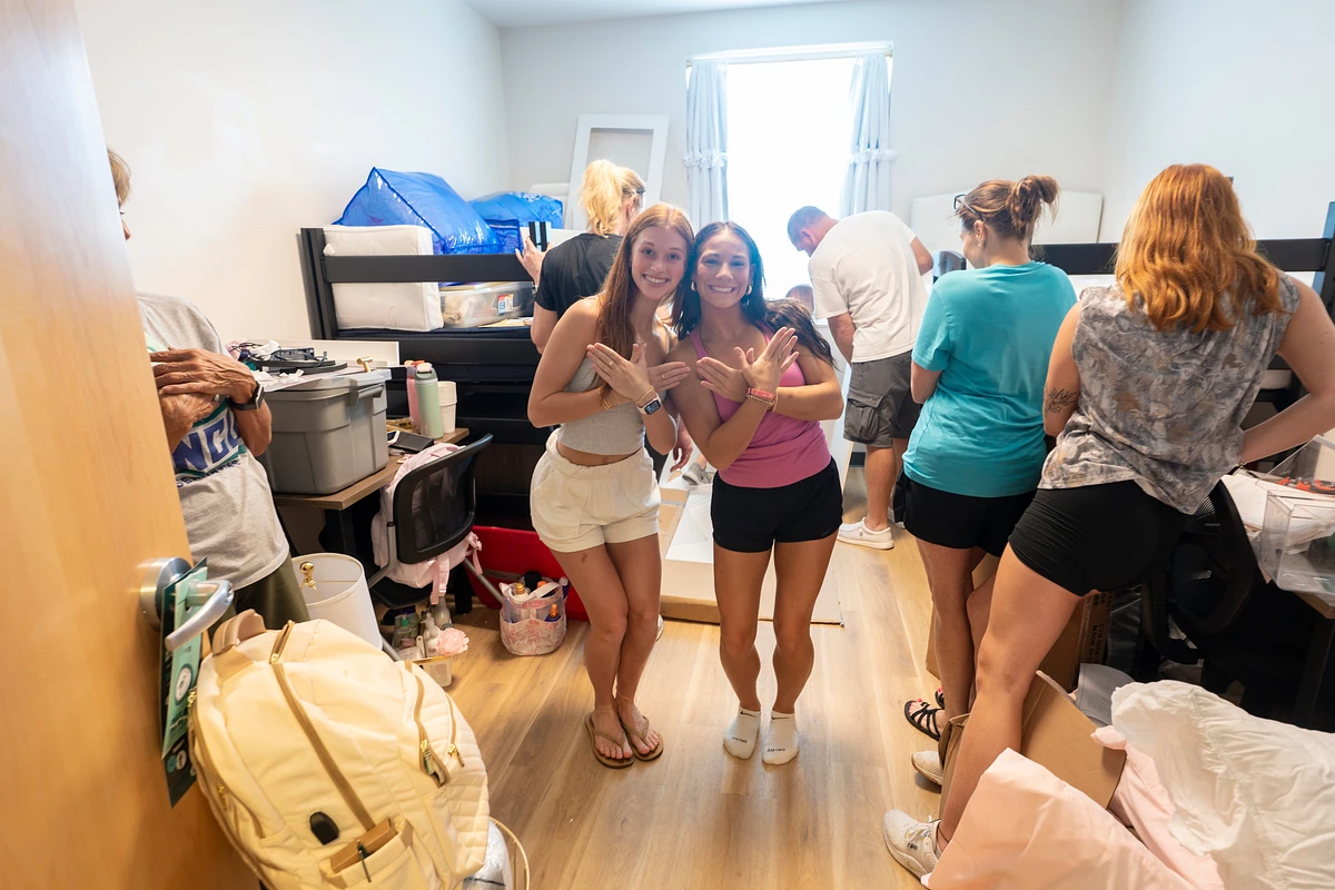 Two young women pose excitedly in a crowded room with several people unpacking and organizing items.